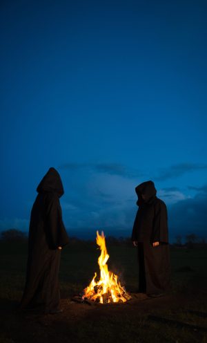 Mysterious, hooded, Pagan figures wearing hoods and cloaks worshiping gods around a fire in a ceremonial ritual at night with copy space